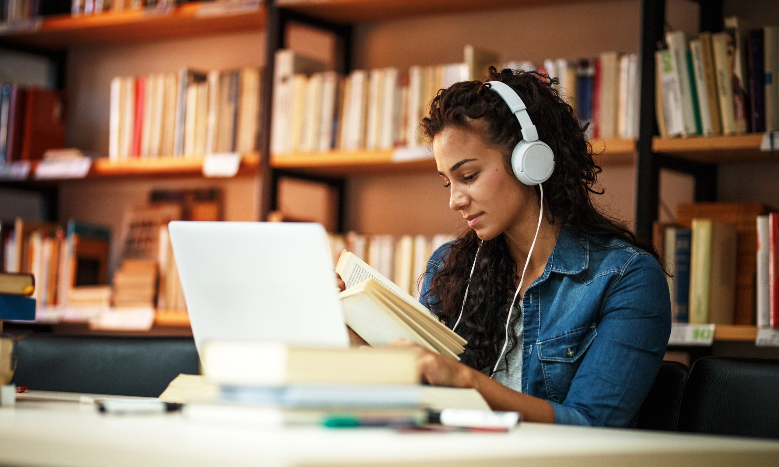 Jeune étudiante lisant un livre avec un casque audio devant son ordinateur dans une bibliothèque.